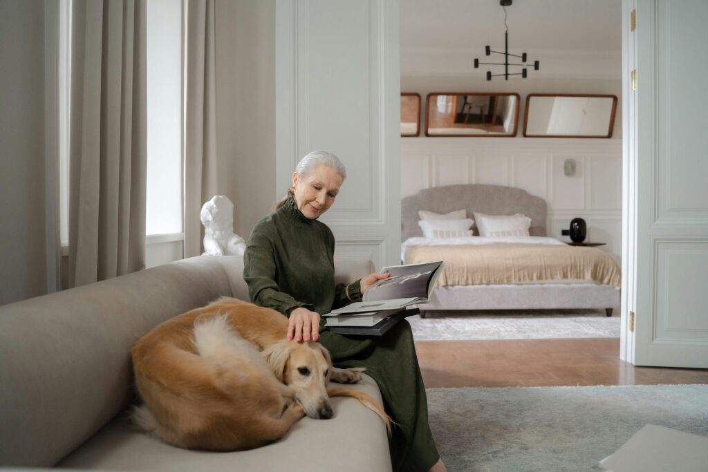Senior woman reading on a sofa while petting her greyhound dog in a cozy living room.