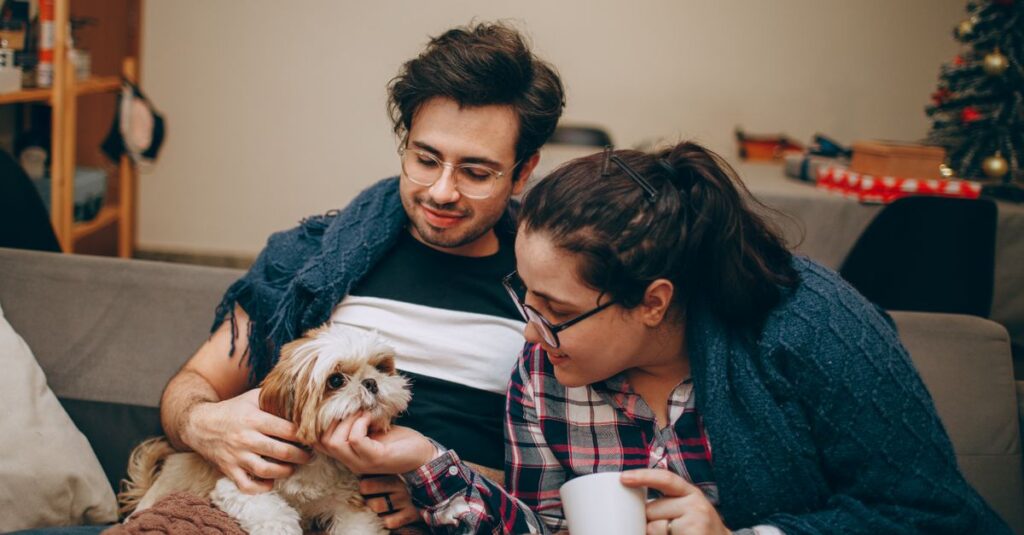 Happy couple relaxing indoors with their cute pet dog during winter, wrapped in blankets.