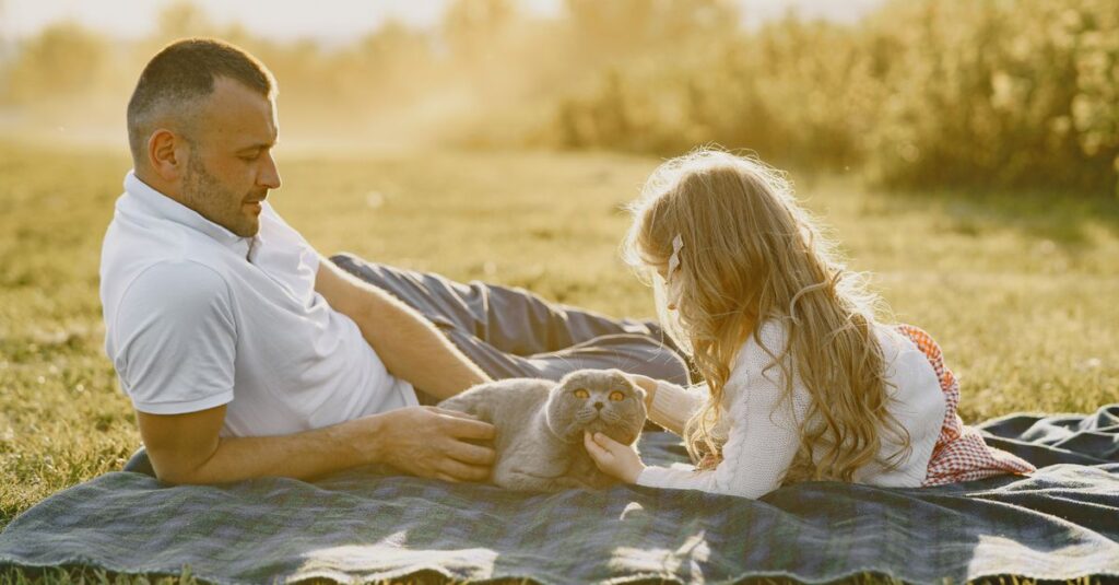 A father and daughter relaxing outdoors with a Scottish Fold cat during a sunny day.