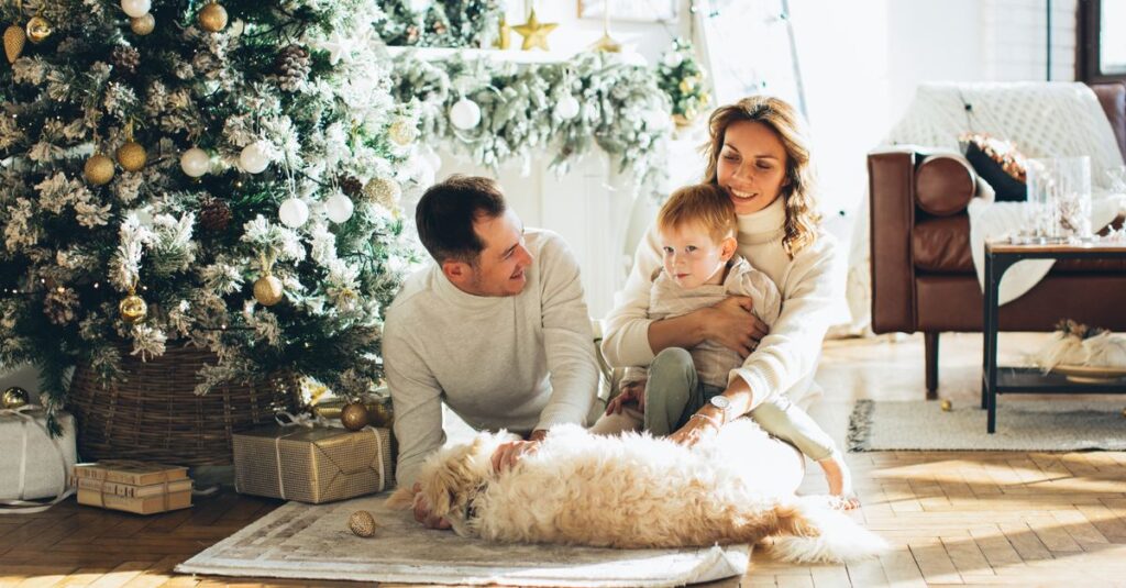 Family of three with a dog sitting by a decorated Christmas tree in a cozy living room.