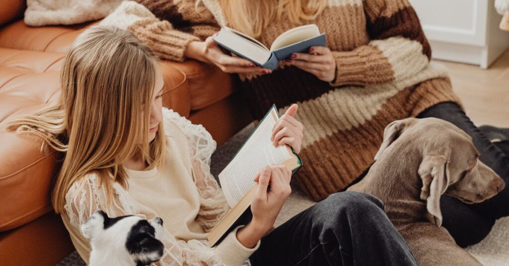 A cozy family scene with mother and daughter reading books together on the sofa with their dogs.