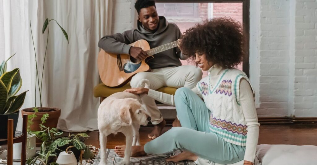 Full body of cheerful African American boyfriend playing guitar near ethnic happy girlfriend with dog on rug in cozy lounge